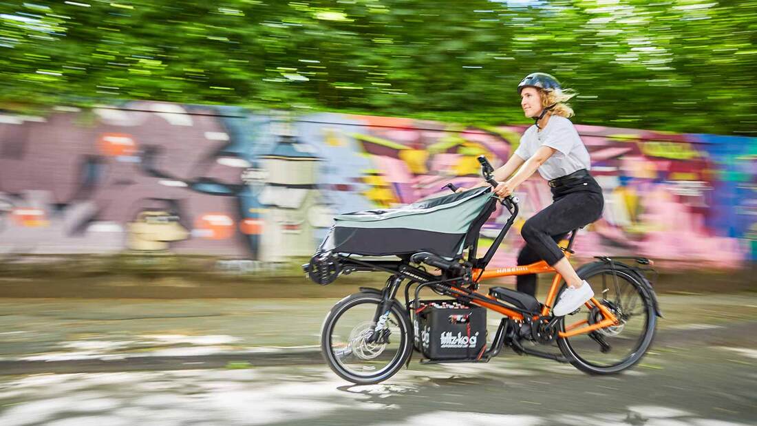 A woman riding a Hase PINO Cargo bike in a city with a graffiti covered wall in the background.