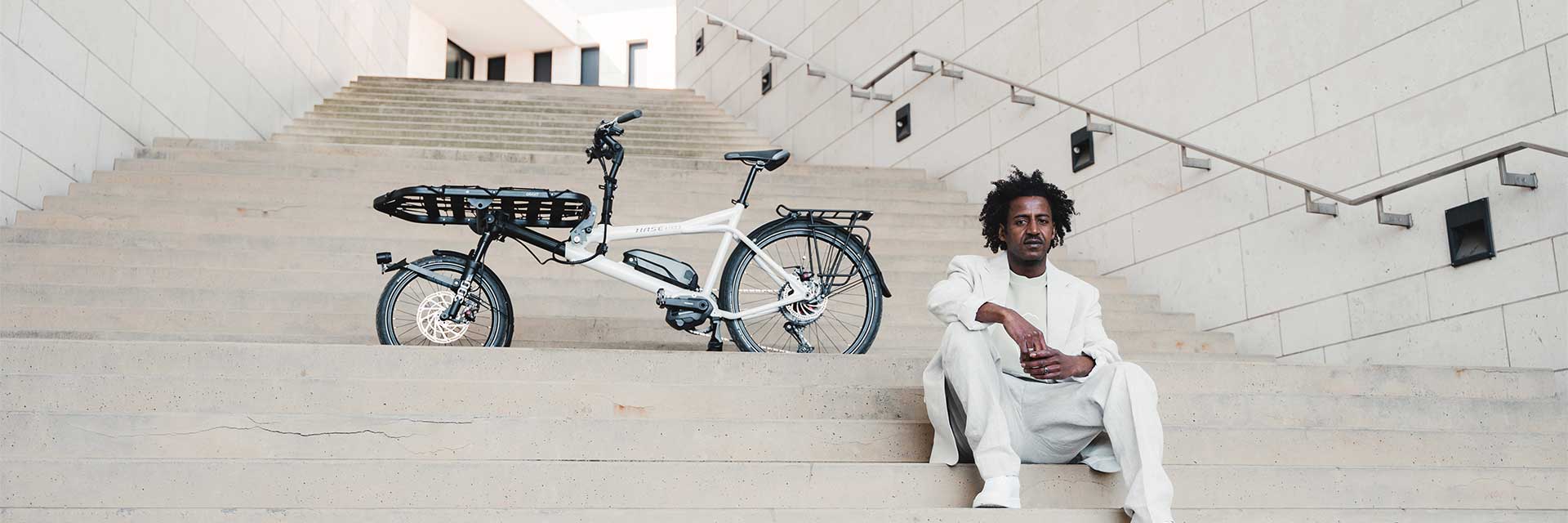 The Hase Gravit City E bike is seen parked on an outdoor stair landing, with a man in a white suit sitting on the steps in front of and below it. The bike is equipped with a cargo board and motor.