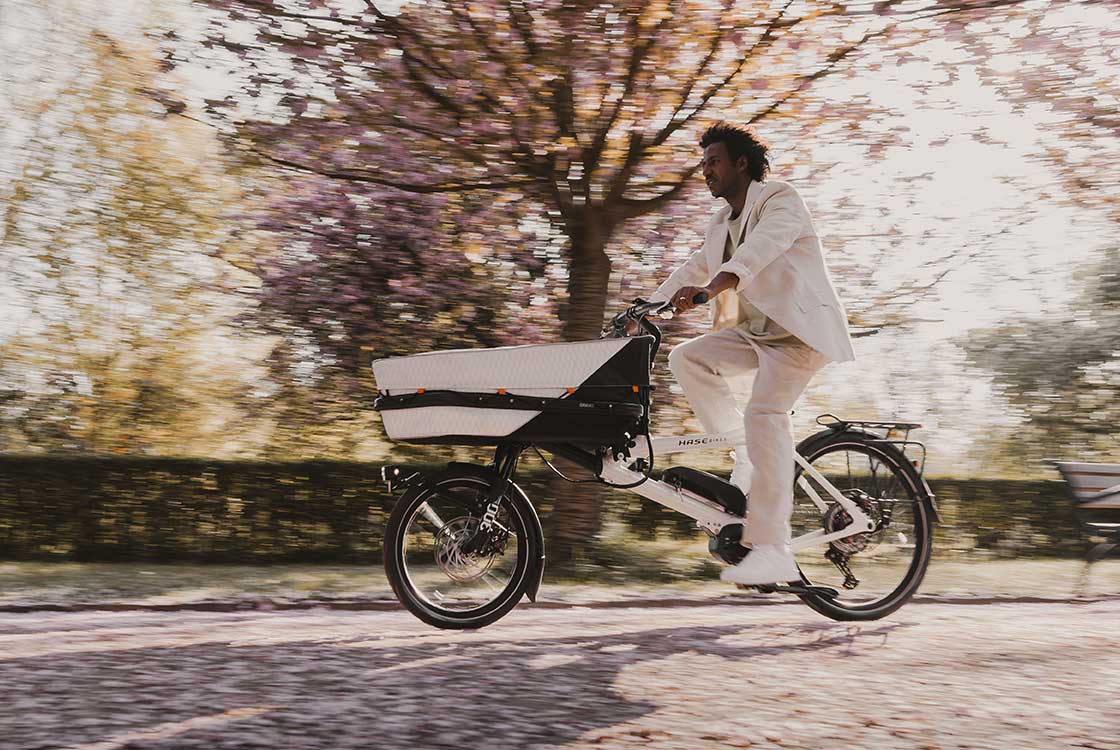 A black man in a white suit rides a GRAVIT CITY E cargo bike in a park. The trees are in bloom behind him.