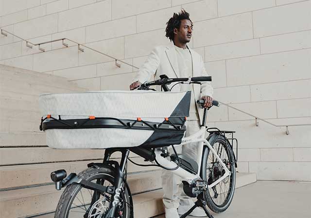 A black man in a white suit stands next to a Hase GRAVIT CITY E cargo bike at the bottom of a flight of stairs. The bike is equipped with a white cargo bag and a Shimano Steps motor.