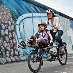 Woman and girl riding a Tandem Pino Allround past a mural